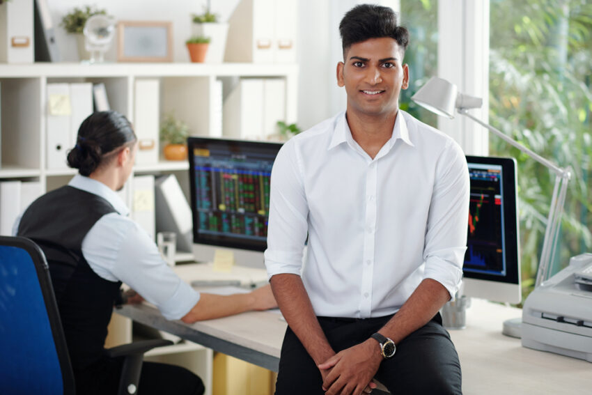 Young man in a white shirt sits on a desk in a busy trading office while a colleague works at a computer with stock charts on the monitor (outdoorsy light).